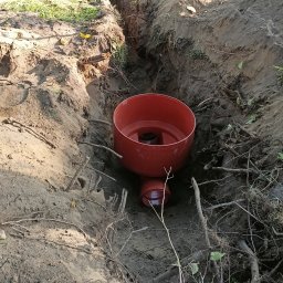 K.P. Digger - Czerwony zbiornik w wykopie ziemnym, prawdopodobnie element instalacji sanitarnej, widoczne korzenie i gałęzie w ziemi.