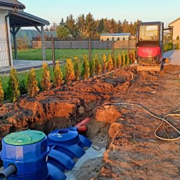 K.P. Digger - Niebieski zbiornik z zieloną pokrywą w wykopie, częściowo wypełnionym wodą. W tle minikoparka Kubota i rząd tuj. Widok na etapie instalacji.