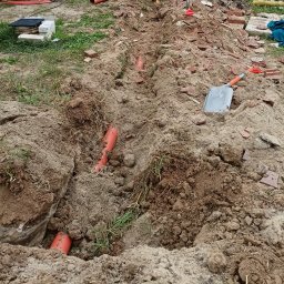 K.P. Digger - Wykop z pomarańczowymi rurami instalacyjnymi w ziemi, widoczne narzędzia (szpadel, łopata) i gruz. Prace ziemne w toku, instalacja podziemna.