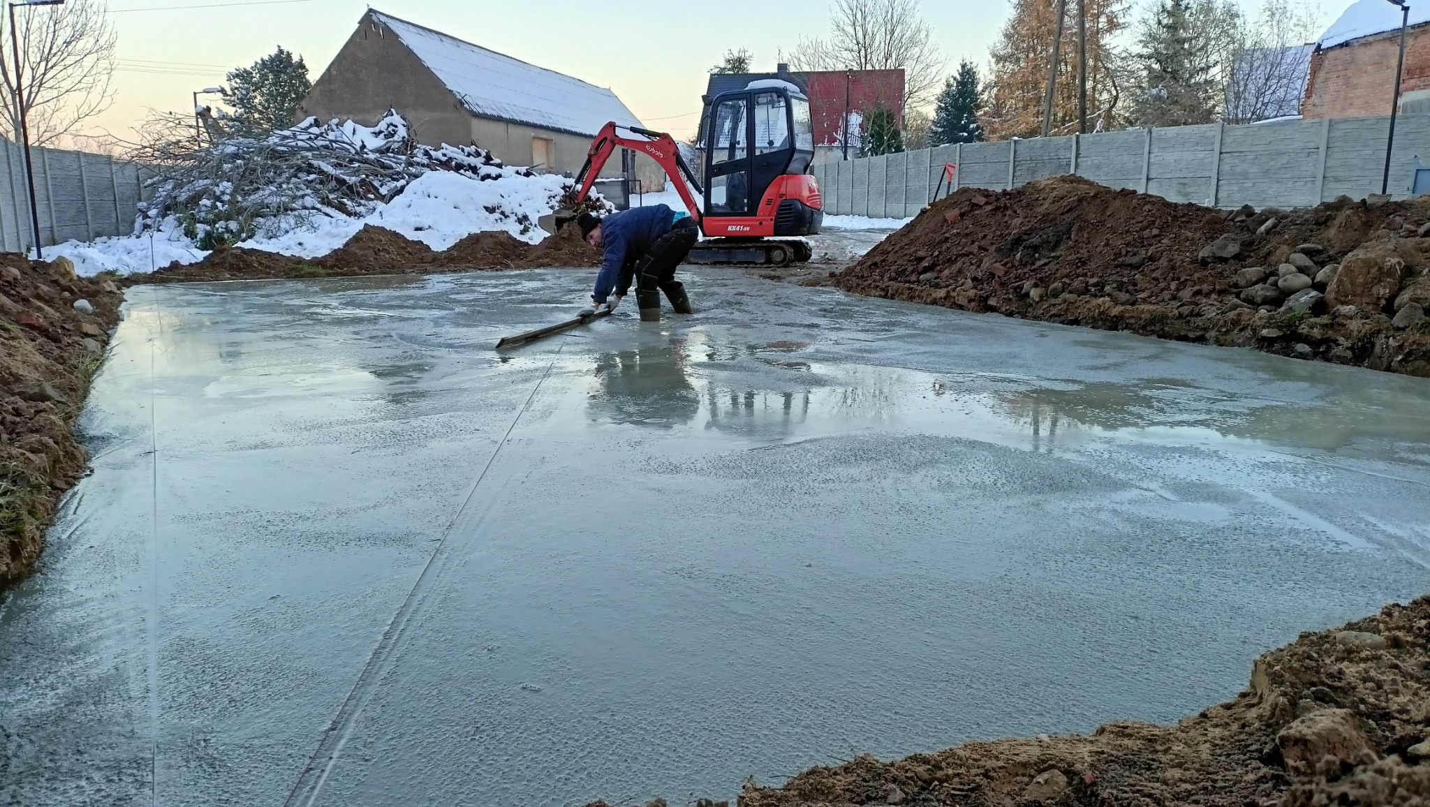 Wyrównywanie wylewki betonowej w wykopie pod fundamenty, na budowie stoi mała koparka, w tle budynek i ogrodzenie. Praca w toku, widoczne narzędzia i materiały.