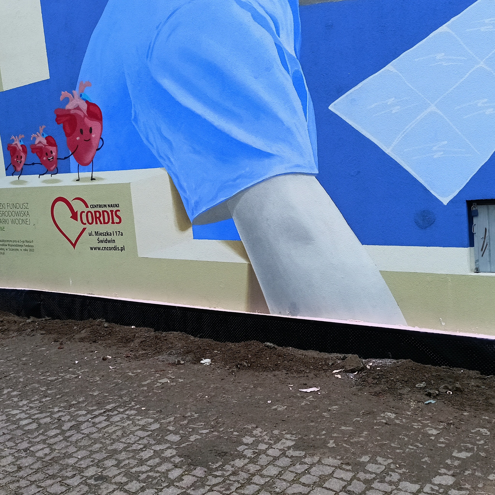 Mural z motywem medycznym: dłoń w niebieskim rękawie, serca z uśmiechem i logo Centrum Nauki Cordis na ścianie budynku. U dołu widoczny chodnik z kostki brukowej.