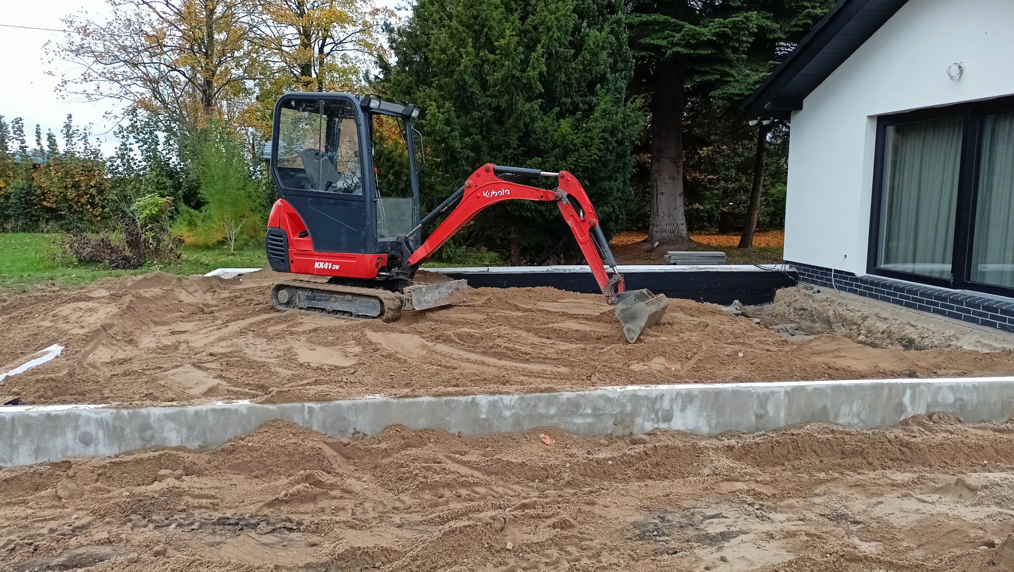 Czerwono-czarna mini koparka Kubota KK41-3U na piasku przy fundamencie domu z czarną podmurówką, w tle drzewa.