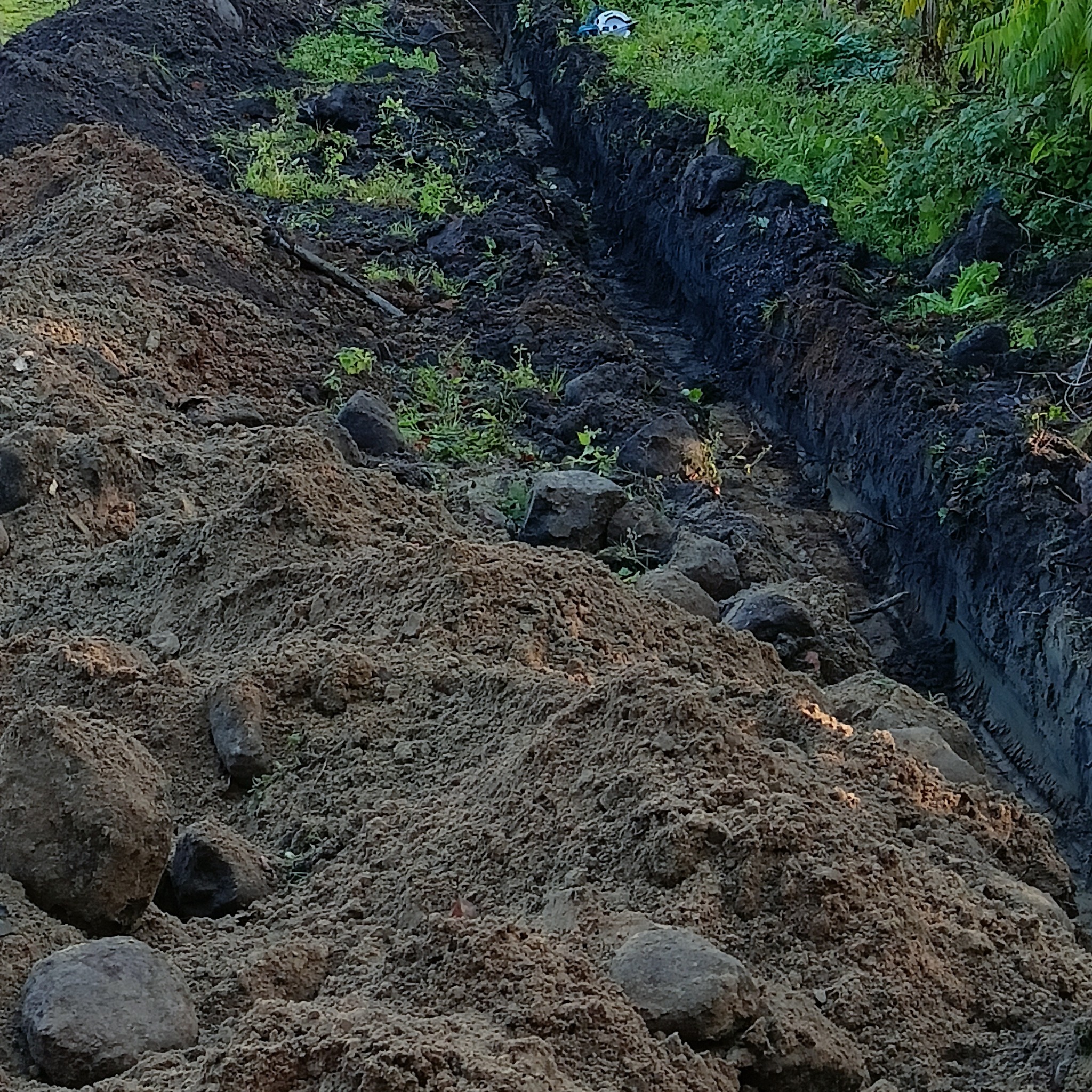 Wykopany rów w ziemi, z hałdą ziemi obok, częściowo porośnięty zielenią. Widoczne odciski opon w wykopie. Kamienie w ziemi.
