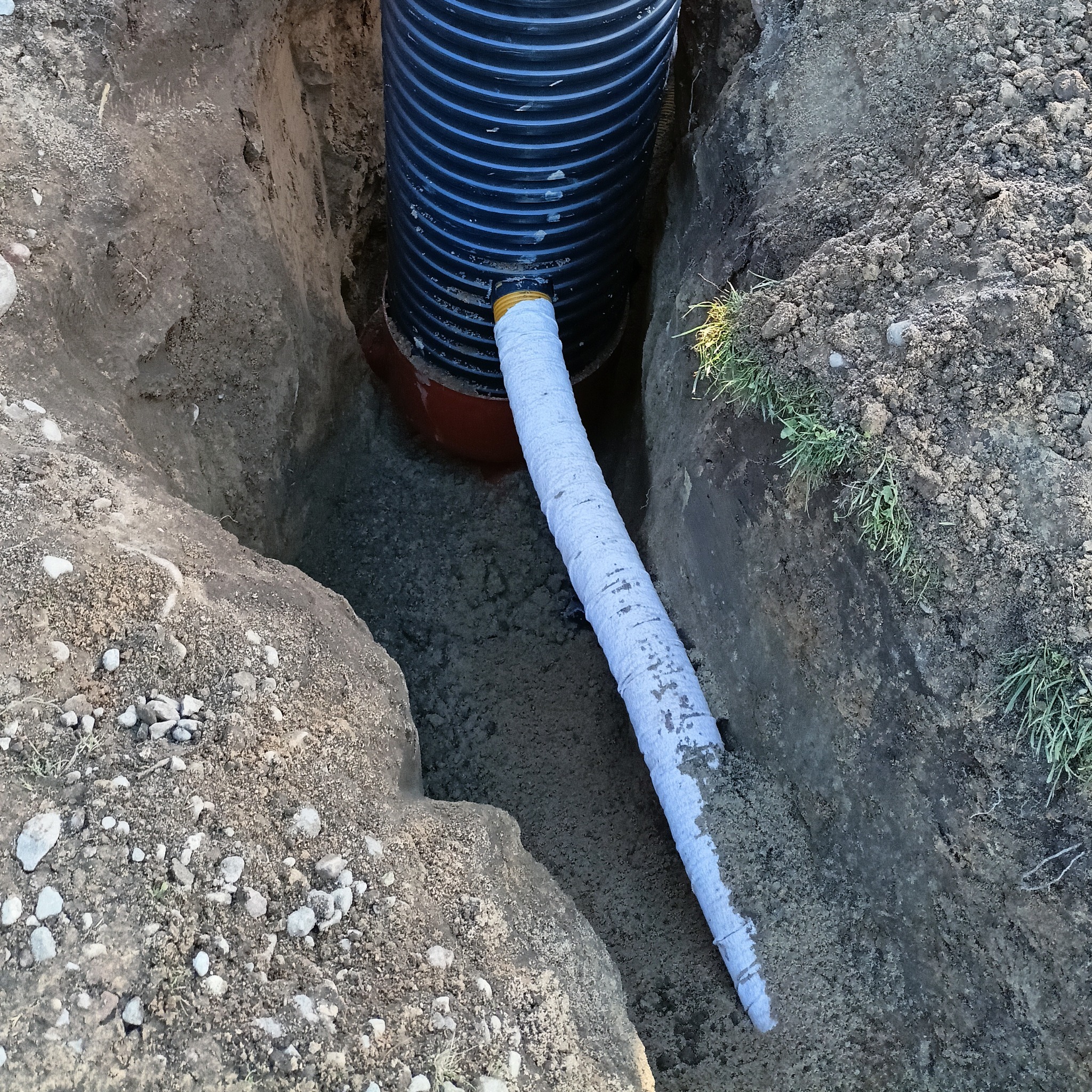 Częściowy widok drenażu w wykopie, czarna rura karbowana z białym owinięciem, ziemia.