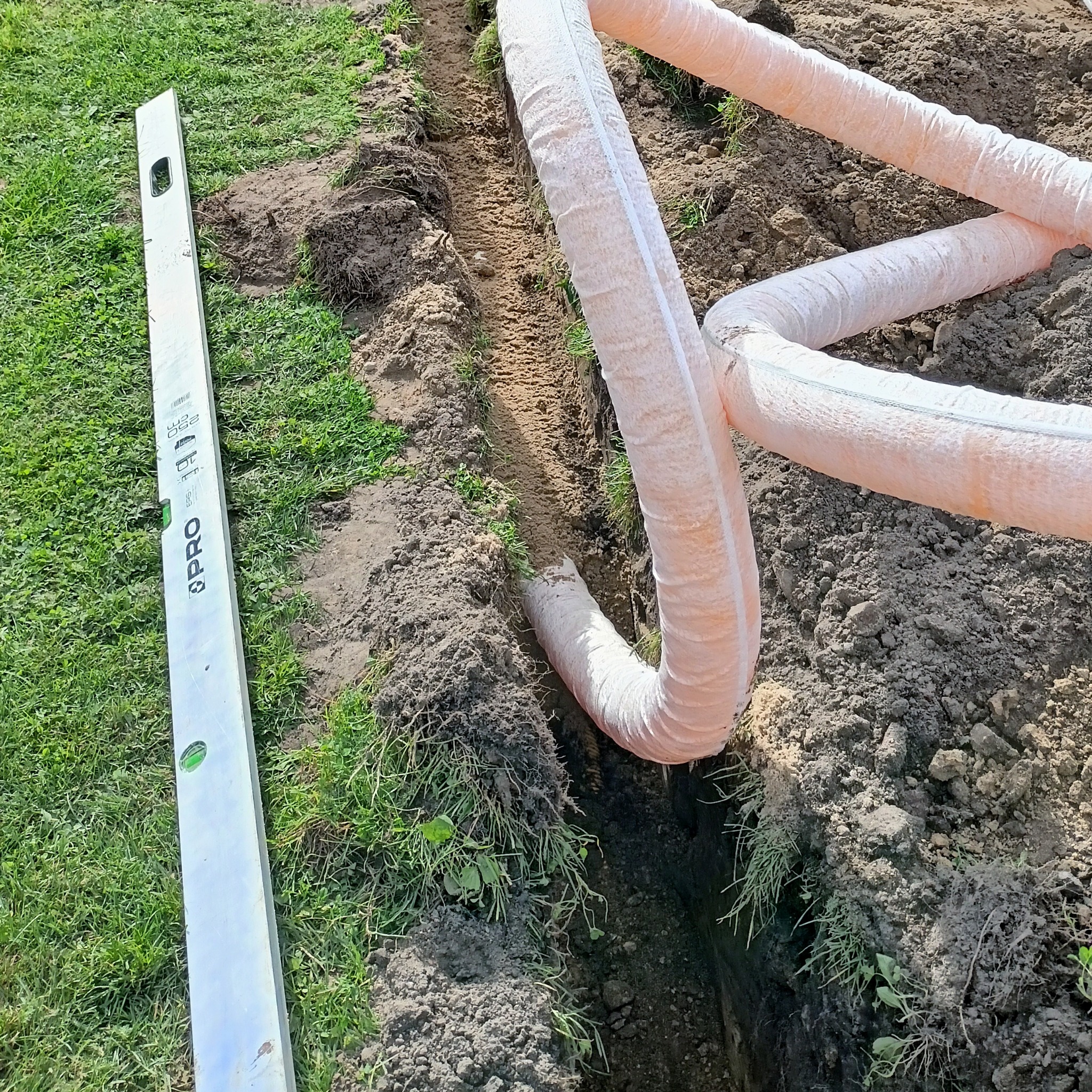 Wykopany rów z ułożonym drenem w pomarańczowym oplocie, obok poziomica na trawie. Widoczna ziemia i trawa. Prace ziemne przy drenażu.