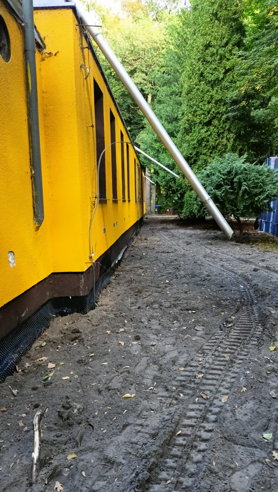 Żółty budynek z izolacją fundamentów i śladami opon w ziemi. Widoczna instalacja rury spustowej. W tle drzewa.