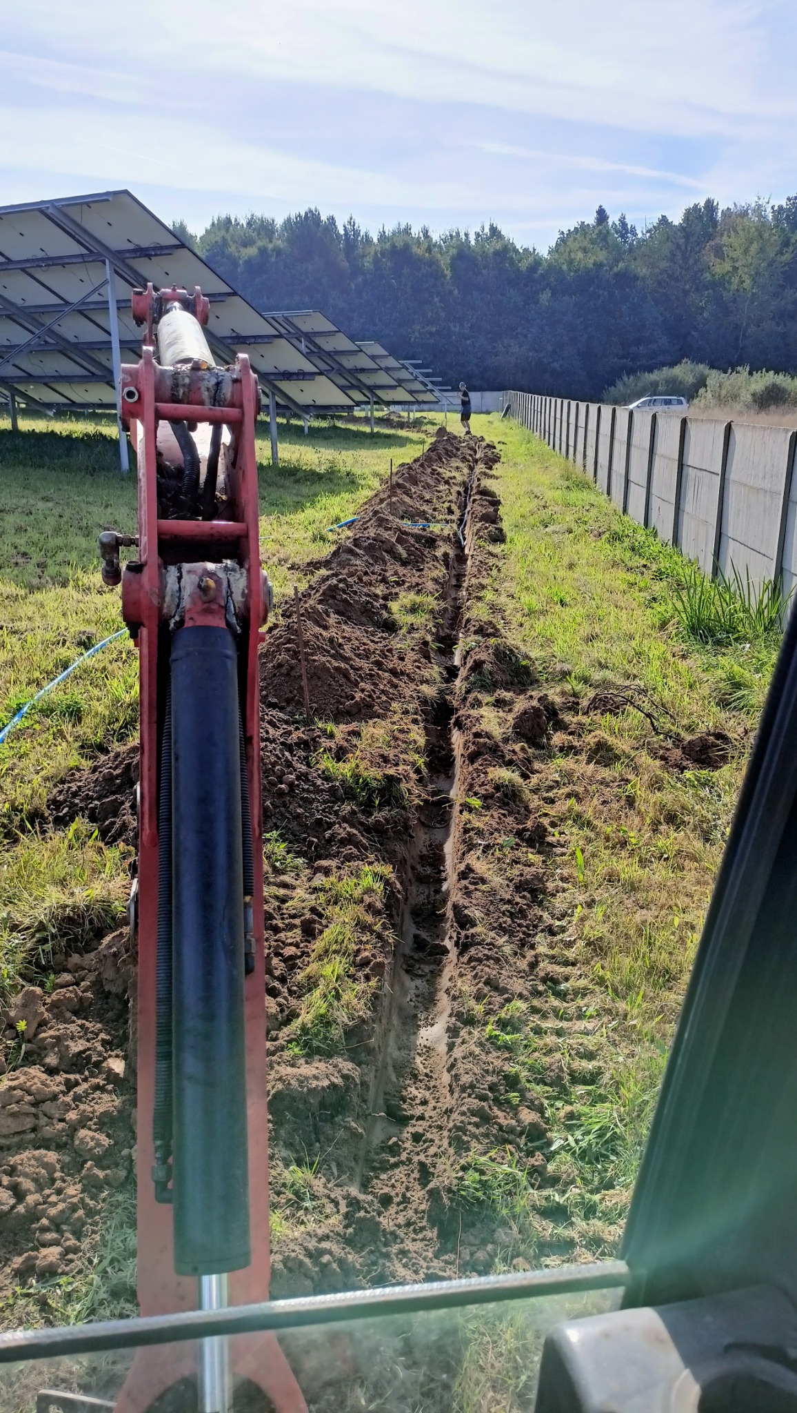 Wykopany rów na zielonym terenie z panelami słonecznymi w tle, widziany z kabiny koparki. Wzdłuż rowu widoczny betonowy płot. Prace ziemne w toku.