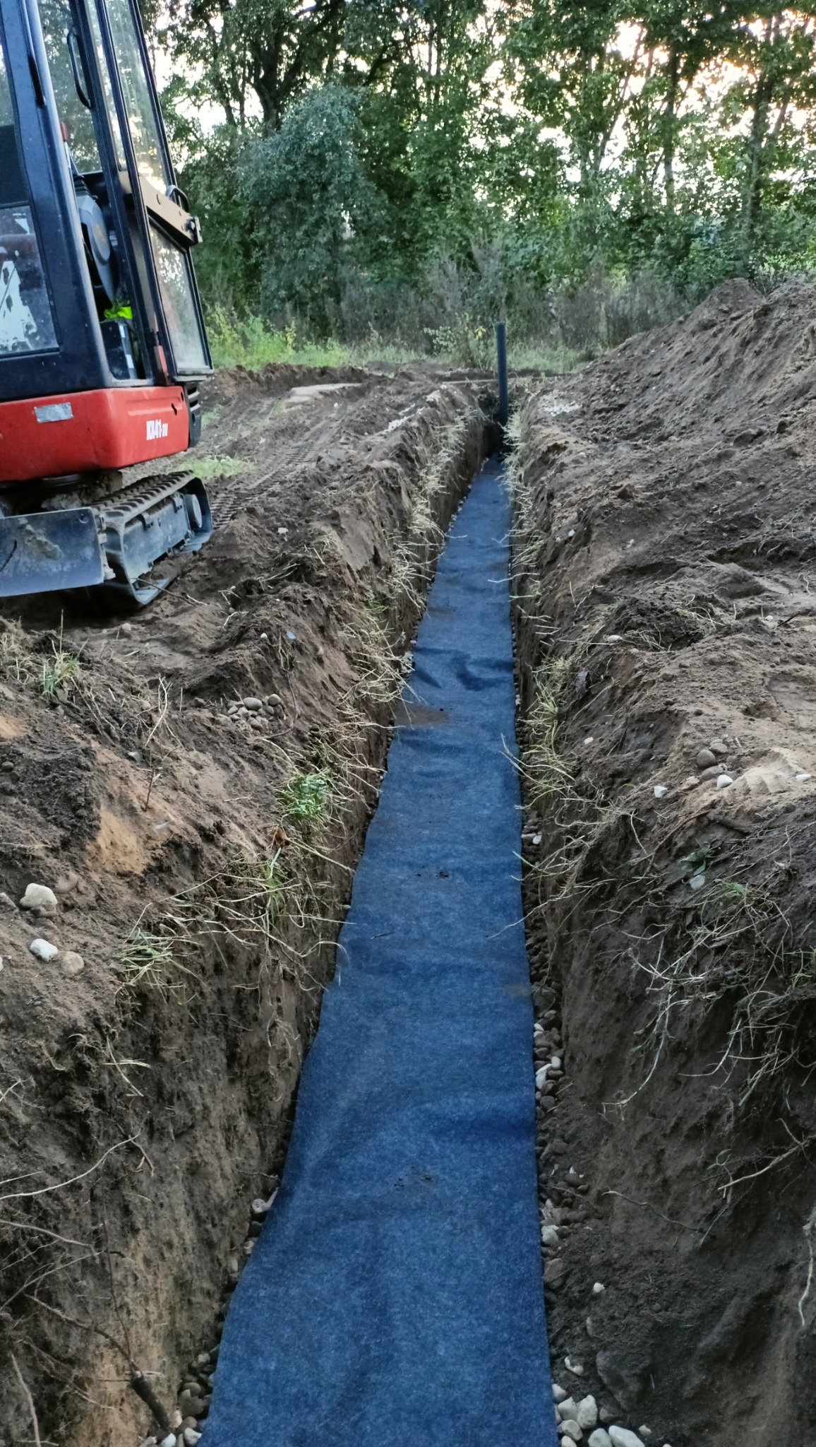 Wykop z wyłożoną geowłókniną, przygotowany pod instalację, z widocznym fragmentem koparki i rurą wystającą z ziemi. Tło stanowią drzewa.