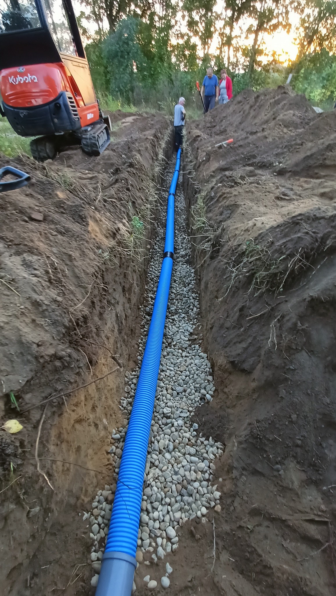 Wykop z niebieską rurą drenażową na żwirowym podłożu, w tle koparka Kubota i ekipa. Widok z góry na wykopaną ziemię, praca w toku.
