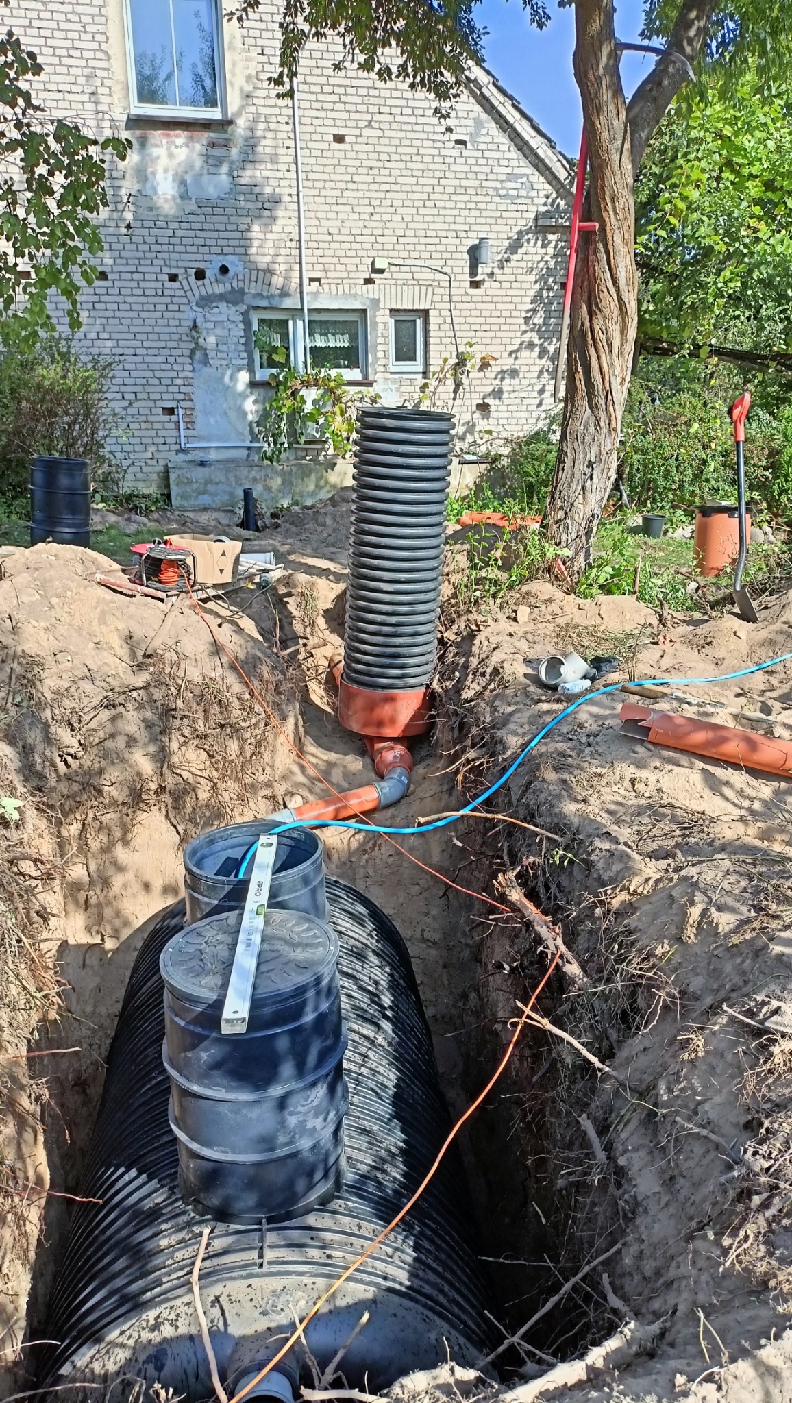 Instalacja zbiornika retencyjnego w wykopie, widoczne rury, studzienka i poziomica. W tle budynek mieszkalny i drzewa. Prace ziemne w toku.