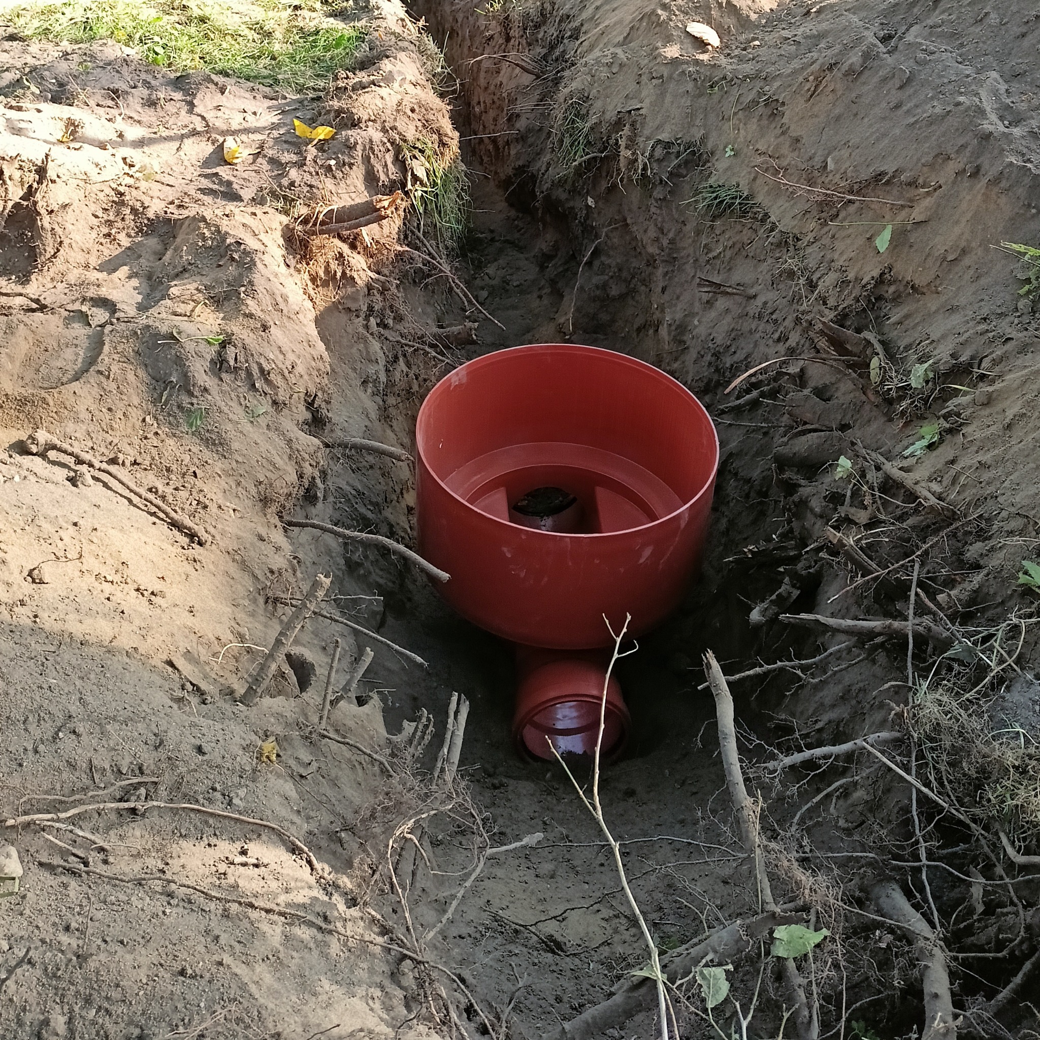 Czerwony zbiornik w wykopie ziemnym, prawdopodobnie element instalacji sanitarnej, widoczne korzenie i gałęzie w ziemi.