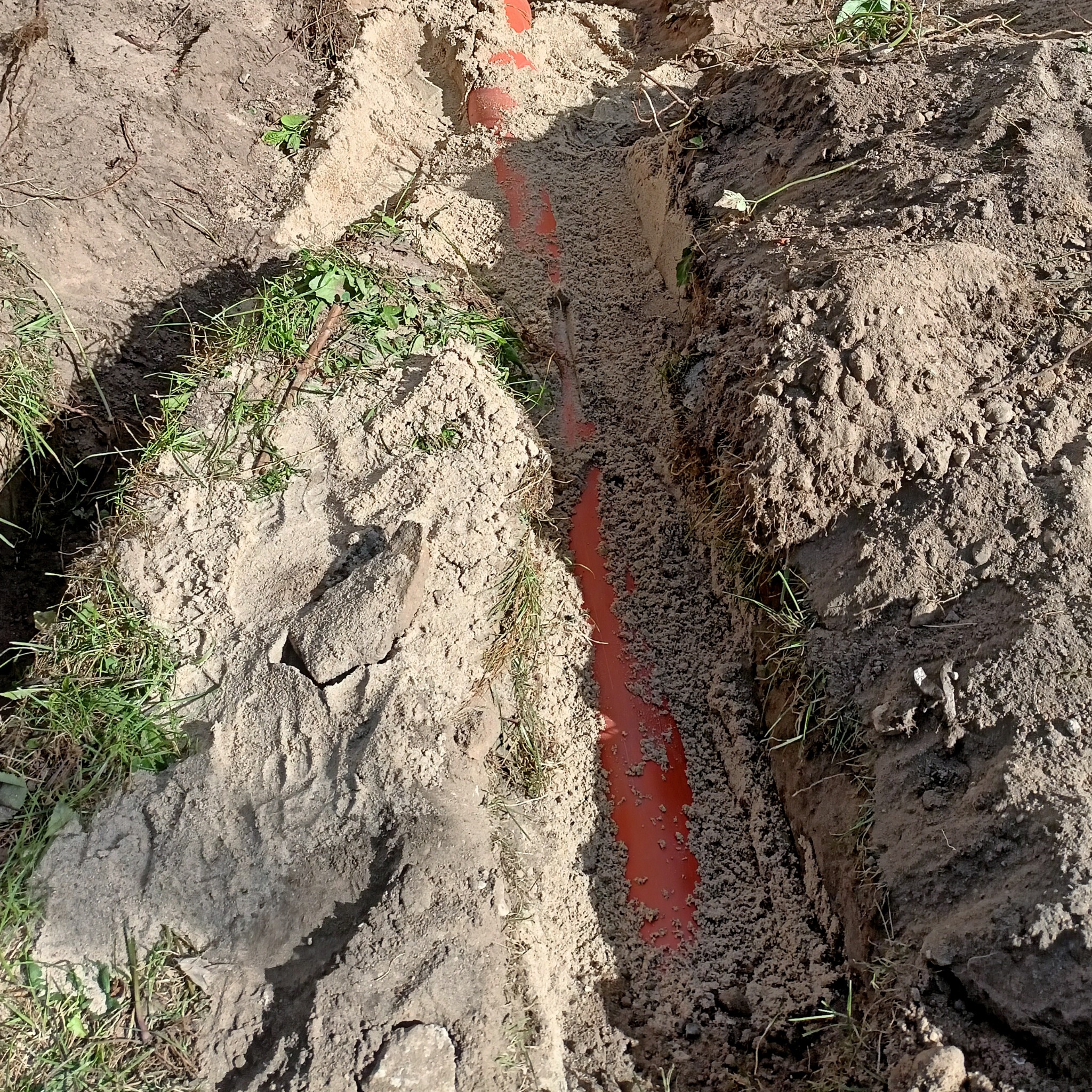 Wykop z pomarańczową rurą w ziemi, częściowo zasypany piaskiem. Widoczne ślady po wykopie i trawa na brzegach wykopu. Rura ułożona w rowie.