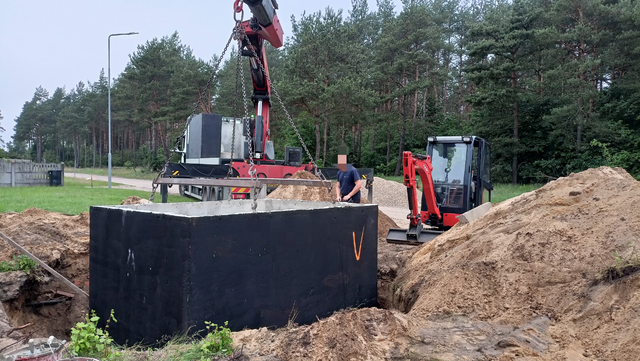 Montaż betonowego szamba: czarny zbiornik opuszczany do wykopu za pomocą dźwigu, obok koparka i pracownik. Widoczne drzewa w tle.
