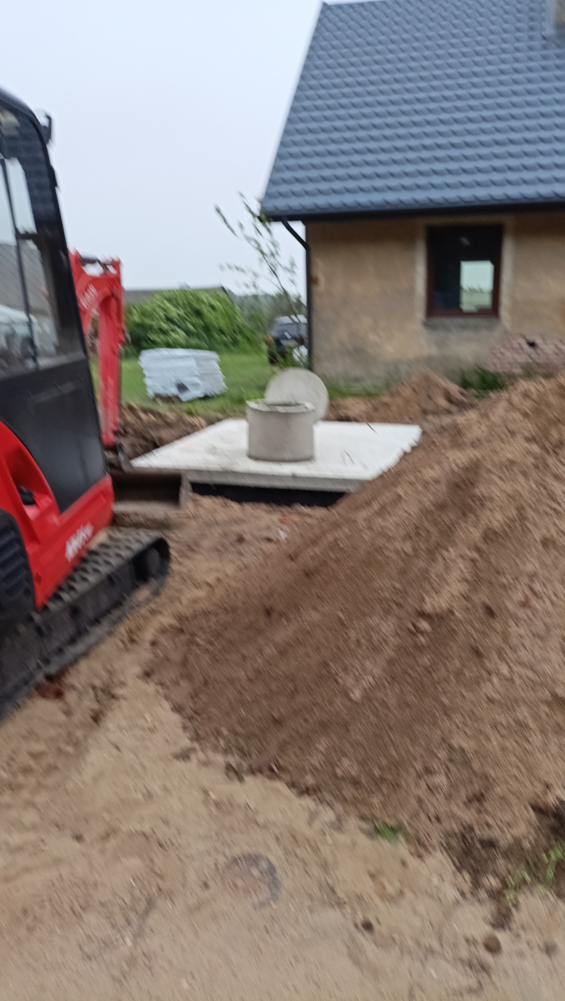 Betonowy zbiornik częściowo zasypany ziemią, widoczny wykop i mała koparka. Prace ziemne w trakcie realizacji, w tle budynek z szarym dachem.