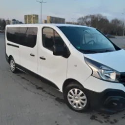 Biały bus Renault Trafic z przyciemnianymi szybami, zaparkowany na szarym brukowanym placu, z widocznym budynkiem mieszkalnym w tle pod błękitnym niebem.