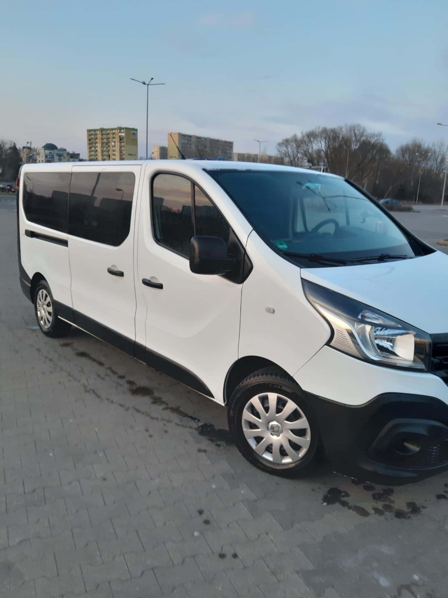 Biały bus Renault Trafic z przyciemnianymi szybami, zaparkowany na szarym brukowanym placu, z widocznym budynkiem mieszkalnym w tle pod błękitnym niebem.