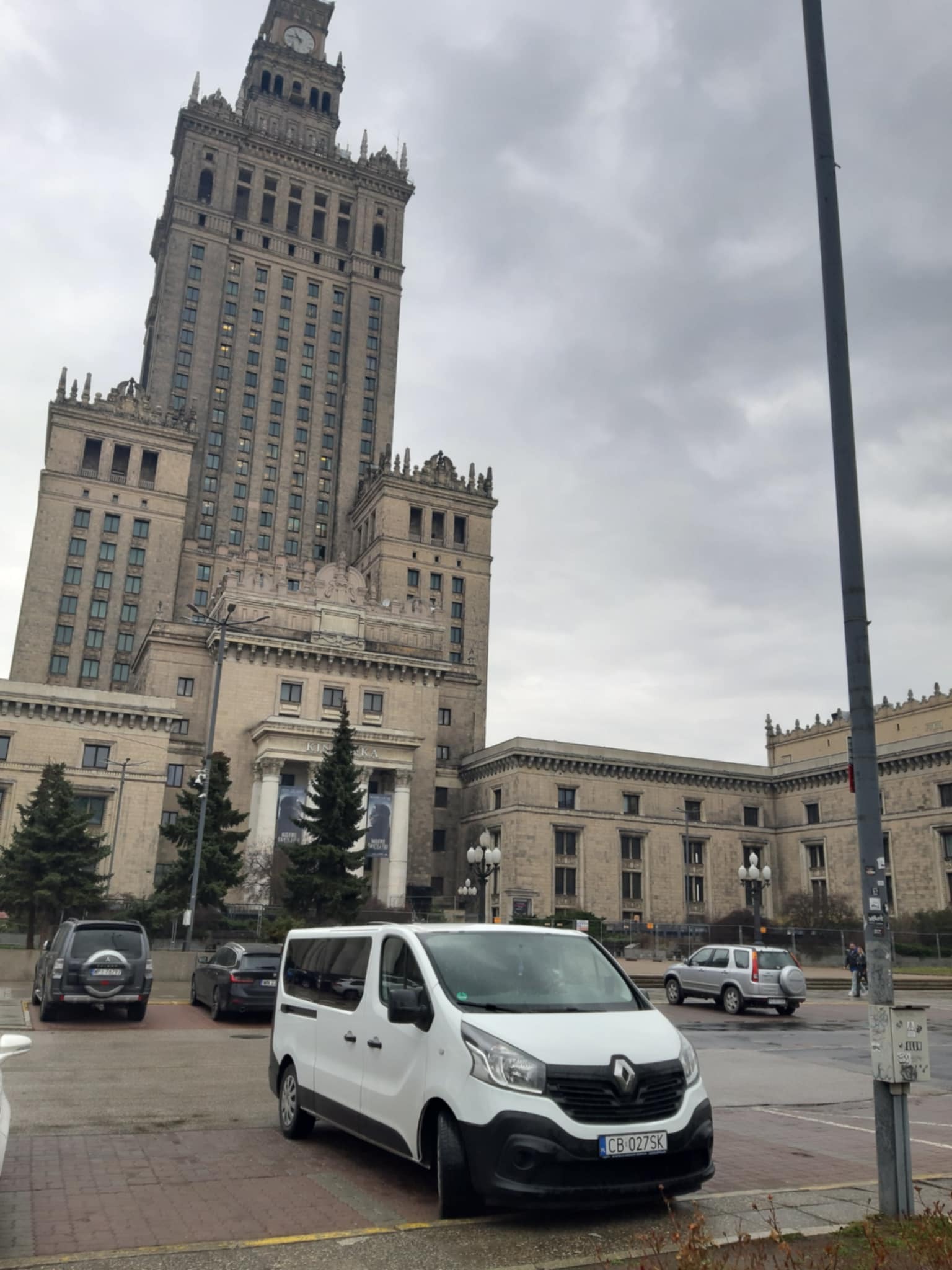 Biały bus Renault Trafic na tle Pałacu Kultury i Nauki w Bydgoszczy, pochmurny dzień.