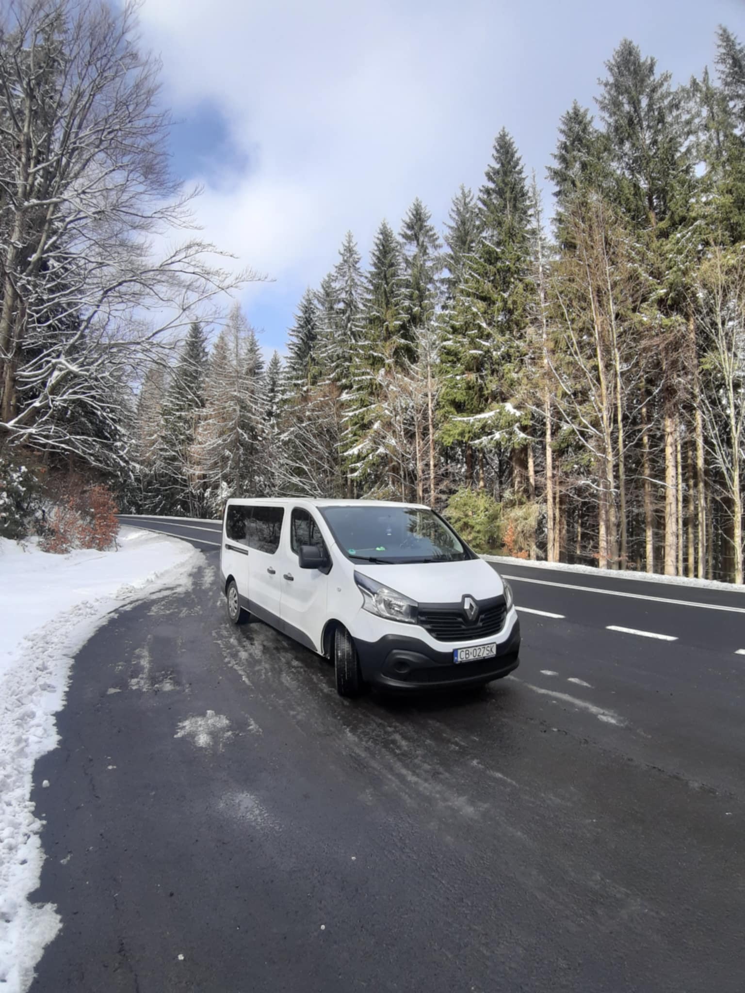 Biały bus Renault Trafic na mokrej, krętej drodze w zimowym krajobrazie, otoczony ośnieżonym lasem iglastym, z widoczną tablicą rejestracyjną.