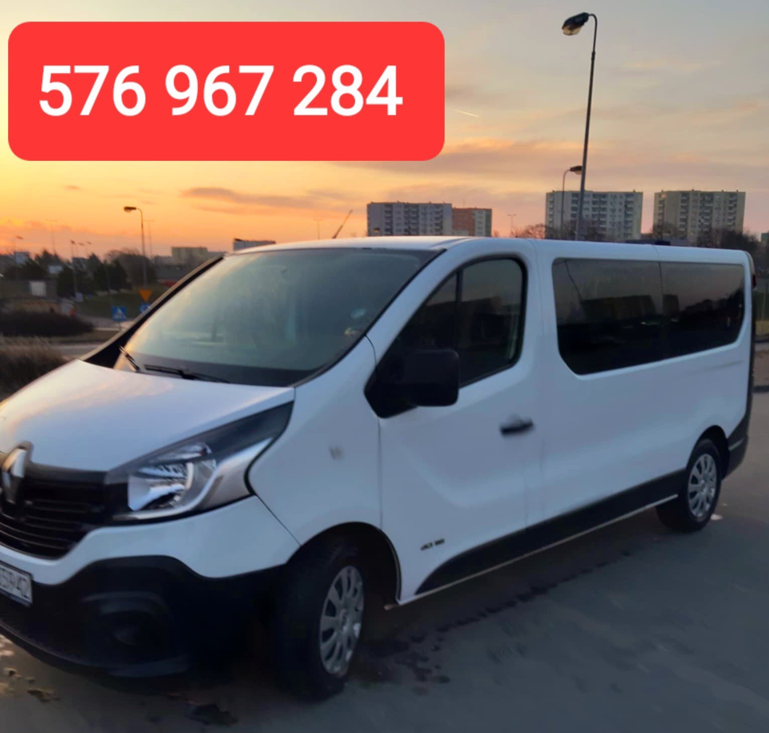 Biały bus Renault Trafic z numerem telefonu na tle zachodzącego słońca i bloków mieszkalnych.