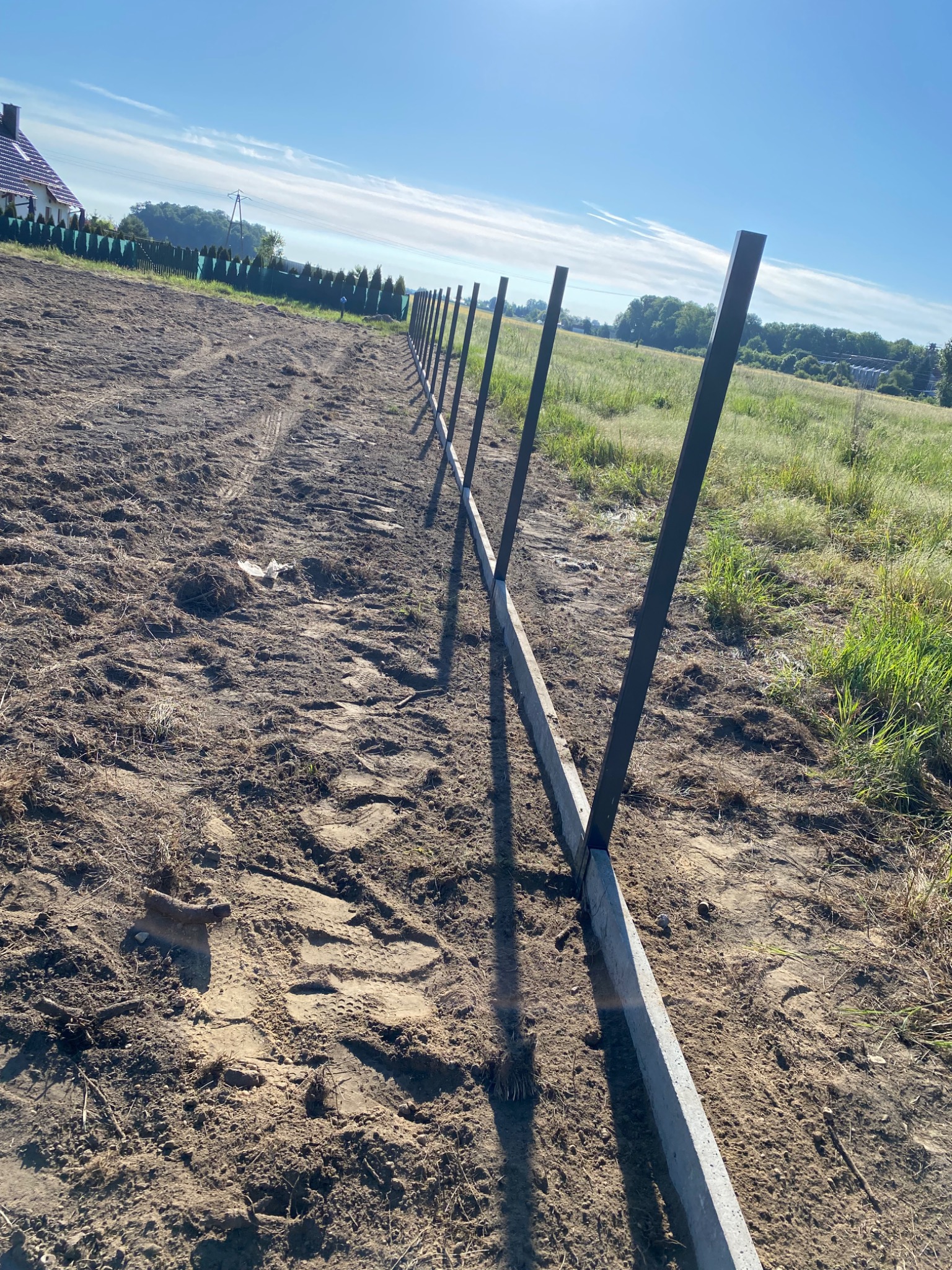 Wyrównany fundament betonowy i słupki z profili stalowych, przygotowane pod montaż ogrodzenia na nierównym terenie z widokiem na dom i pola w tle.