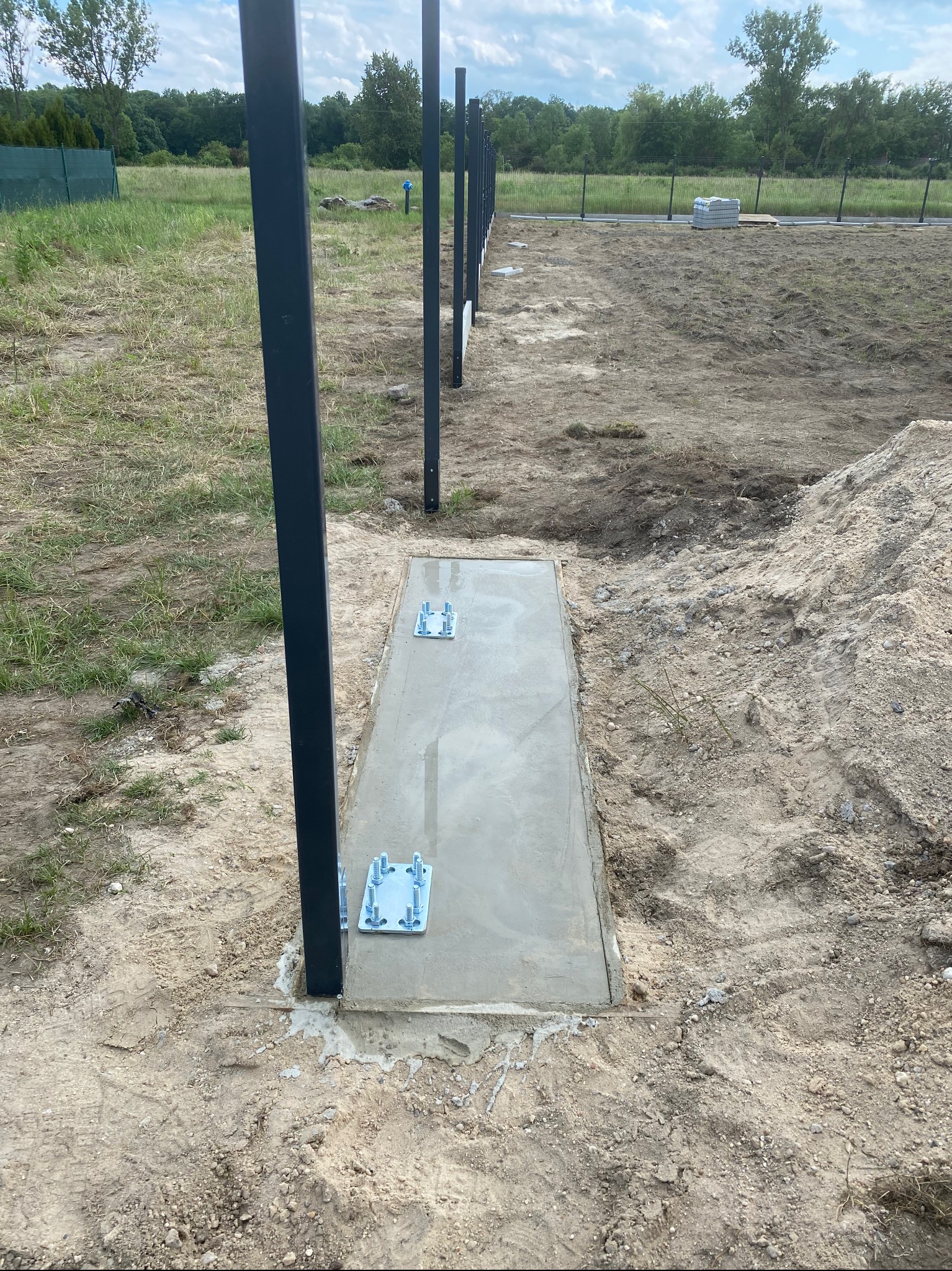 Wylewka betonowa pod słupki ogrodzeniowe z zamocowanymi metalowymi podstawami, widok na rząd słupków w terenie niezabudowanym.