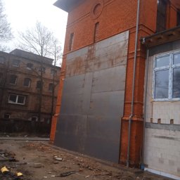 PIOTR TREPCZYK USŁUGI SPAWALNICZE - Duża, szara, metalowa konstrukcja przylegająca do ceglanego budynku, prawdopodobnie zabezpieczenie lub element konstrukcyjny w trakcie budowy, widoczne gruz i materiały budowlane na ziemi.