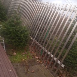 PIOTR TREPCZYK USŁUGI SPAWALNICZE - Długa, nowoczesna ogrodowa konstrukcja z metalowych, prostokątnych profili, ustawionych ukośnie, widoczna z perspektywy pod kątem, z młodymi drzewkami iglastymi w tle i drewnianym podłożem w dolnej...