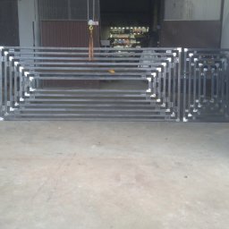 PIOTR TREPCZYK USŁUGI SPAWALNICZE - Metalowa konstrukcja bramy wjazdowej z kwadratowym wzorem, w trakcie produkcji w warsztacie, widoczne spawane łączenia, surowy beton na podłodze.