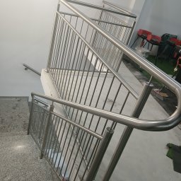 PIOTR TREPCZYK USŁUGI SPAWALNICZE - Widok z góry na schody z balustradą ze stali nierdzewnej o minimalistycznym designie, z widocznym fragmentem szarej podłogi i ściany, w tle częściowo widoczne grille na kółkach.
