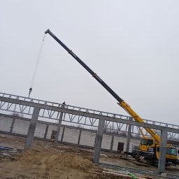 PIOTR TREPCZYK USŁUGI SPAWALNICZE - Żółty dźwig montuje stalową konstrukcję dachu na placu budowy hali z prefabrykatów betonowych w pochmurny dzień.