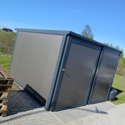 PIOTR TREPCZYK USŁUGI SPAWALNICZE - Nowoczesny garaż blaszany z dwoma segmentowymi bramami w kolorze grafitowym, umieszczony na kostce brukowej, na tle zieleni i błękitnego nieba.