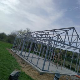 PIOTR TREPCZYK USŁUGI SPAWALNICZE - Konstrukcja metalowa w trakcie montażu, widoczny pracownik na drabinie, zielone otoczenie, pochmurne niebo.  Metalowa rama budynku na fundamencie.