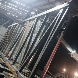 PIOTR TREPCZYK USŁUGI SPAWALNICZE - Wnętrze hali produkcyjnej z surową, metalową konstrukcją w trakcie montażu. Widoczny wózek widłowy i elementy spawarki. Nocne oświetlenie.