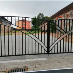 Czarna, metalowa brama wjazdowa z ozdobnymi łukami na tle budynków i szarej kostki brukowej. Widoczny zamek na bramie.