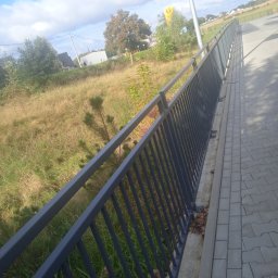 PIOTR TREPCZYK USŁUGI SPAWALNICZE - Długa, szara barierka z metalowych prętów, zamontowana wzdłuż chodnika. W tle trawa i drzewa pod błękitnym niebem. Widok z perspektywy lekko z góry.