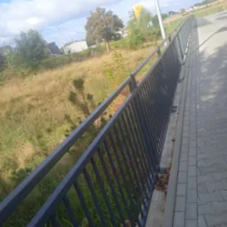 Długa, szara barierka z metalowych prętów, zamontowana wzdłuż chodnika. W tle trawa i drzewa pod błękitnym niebem. Widok z perspektywy lekko z góry.