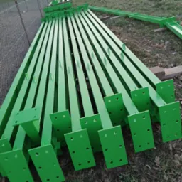 Stalowe, pomalowane na zielono słupki z podstawami z otworami, ułożone równolegle na ziemi, przygotowane do montażu. Widoczna siatka ogrodzeniowa w tle.