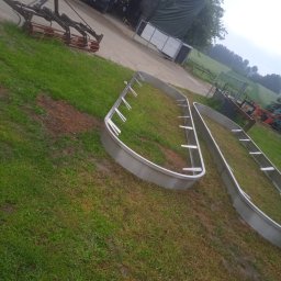 PIOTR TREPCZYK USŁUGI SPAWALNICZE - Dwie metalowe, owalne konstrukcje z uchwytami, leżące na trawie w wiejskim otoczeniu. W tle widoczne maszyny rolnicze i budynek gospodarczy.