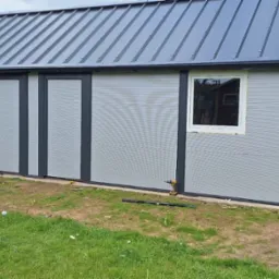 Szary garaż blaszany z metalowym dachem, widoczne narzędzia i skrzynka narzędziowa na trawie. Ukończona konstrukcja z zamontowanymi drzwiami i oknami.