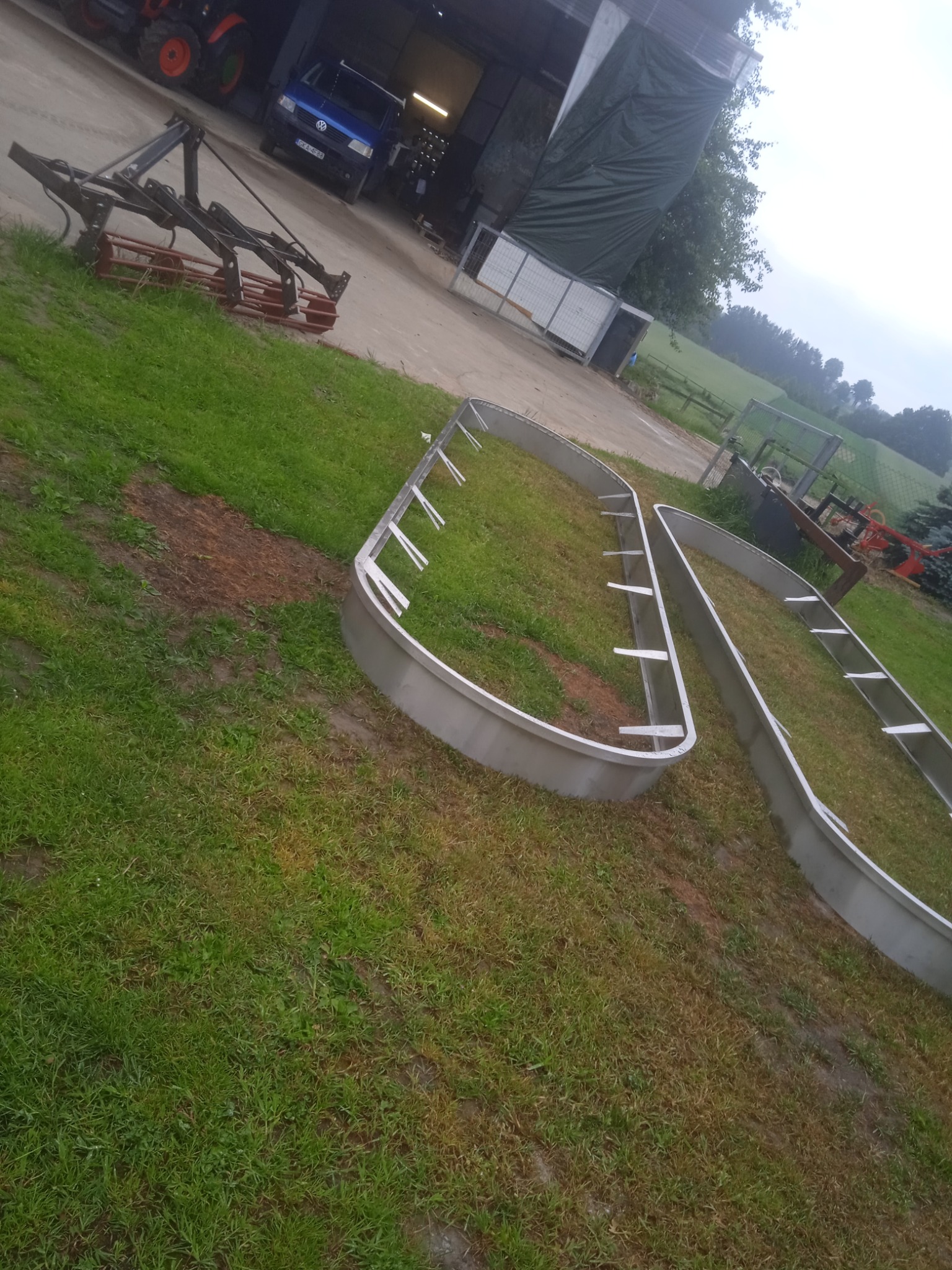Dwa metalowe, owalne obramowania, prawdopodobnie do pergoli, leżące na trawie na tle wiejskiego krajobrazu z budynkiem gospodarczym, traktorem i samochodem.