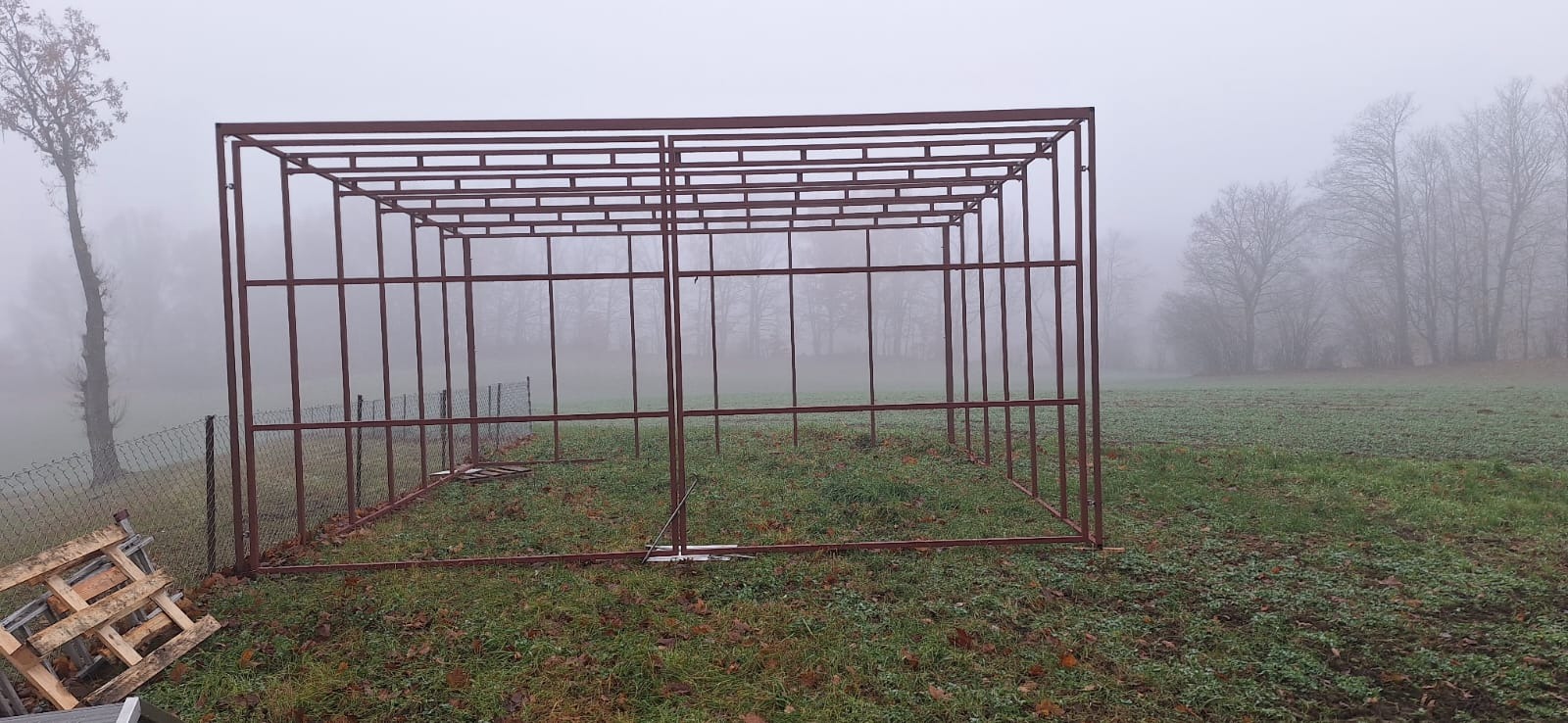 Szkielet metalowej konstrukcji w trakcie budowy na zewnątrz w mglisty dzień, widoczne palety obok ogrodzenia z siatki.