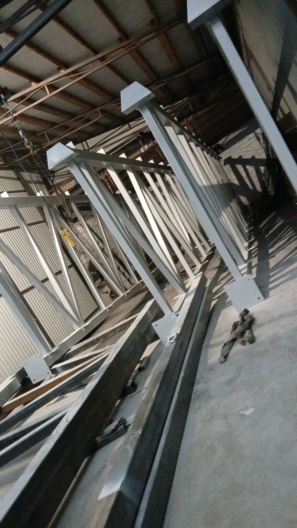 Konstrukcja stalowa w trakcie montażu w hali produkcyjnej, widoczne elementy kratownicy i słupy pomalowane na szaro, leżące na betonowej posadzce w słabym oświetleniu.