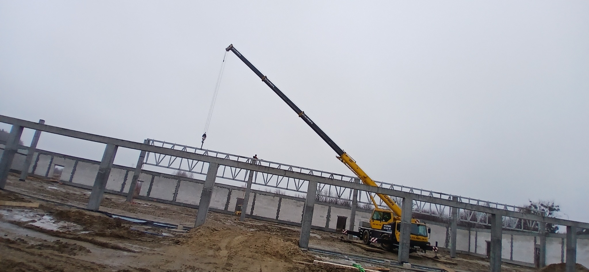 Żółty dźwig montuje stalową konstrukcję dachu na placu budowy hali z prefabrykatów betonowych w pochmurny dzień.