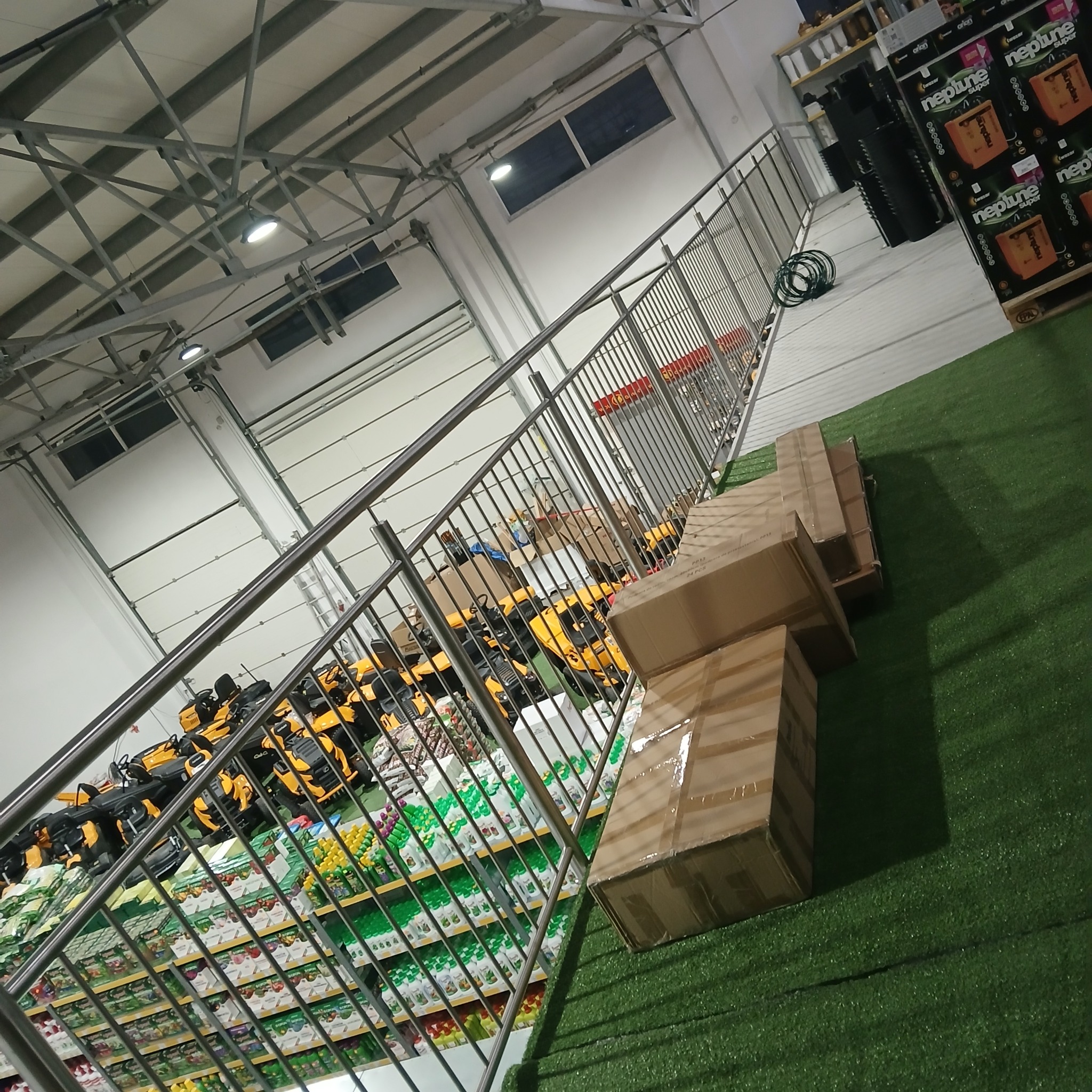 Wnętrze hali magazynowej z metalową balustradą, regałami wypełnionymi towarem i składowanymi kartonami na sztucznej trawie.