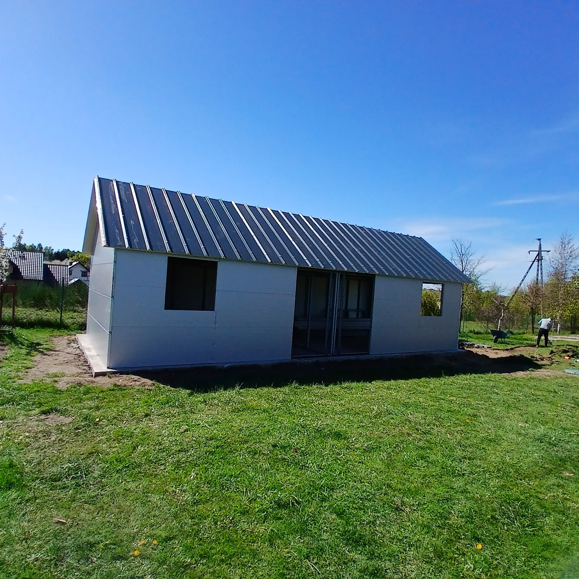 Nowy garaż blaszany z metalowym dachem, częściowo otwarty, na trawniku w słoneczny dzień. Widoczny fundament i otwory okienne.