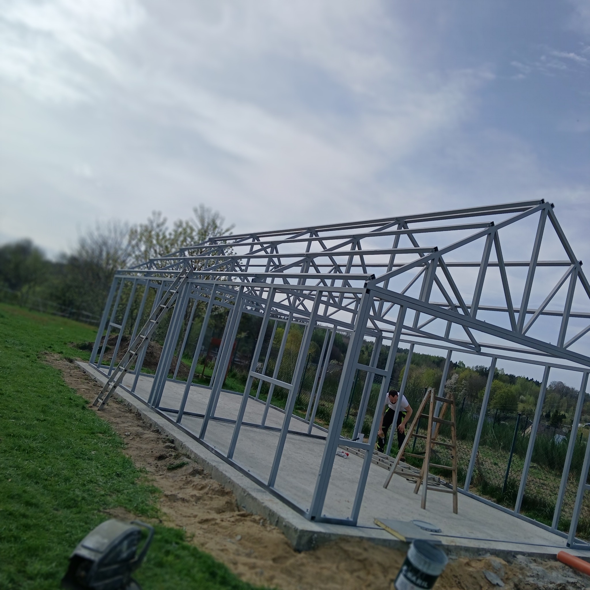 Konstrukcja metalowa w trakcie montażu, widoczny pracownik na drabinie, zielone otoczenie, pochmurne niebo.  Metalowa rama budynku na fundamencie.