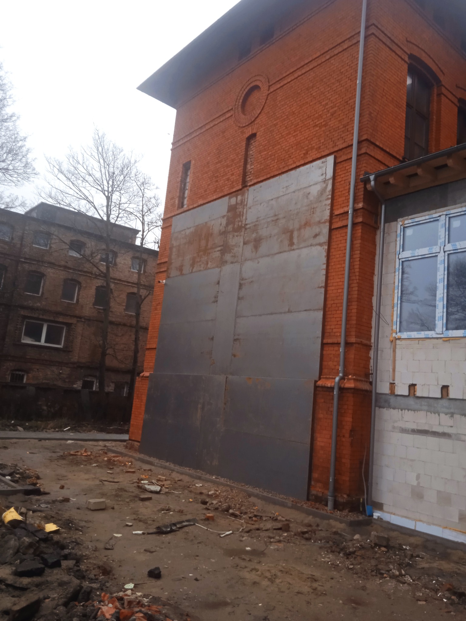 Duża, metalowa płyta zakrywająca wejście do budynku z czerwonej cegły. Obok budynek w trakcie budowy z widocznymi oknami. Ziemia usiana gruzem.