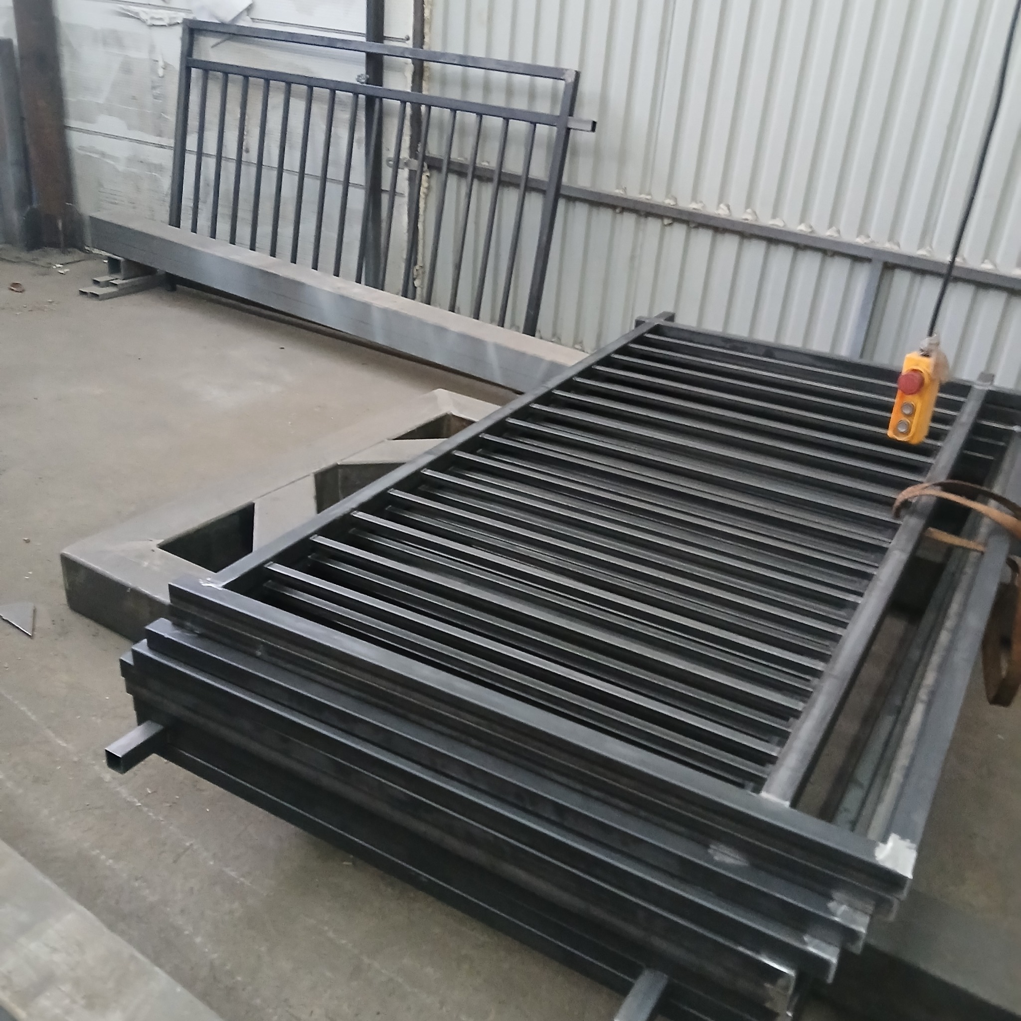 Stos metalowych, spawanych elementów konstrukcyjnych, prawdopodobnie balustrady lub fragmenty bramy, w warsztacie. Widoczny suwnica i metalowe profile.