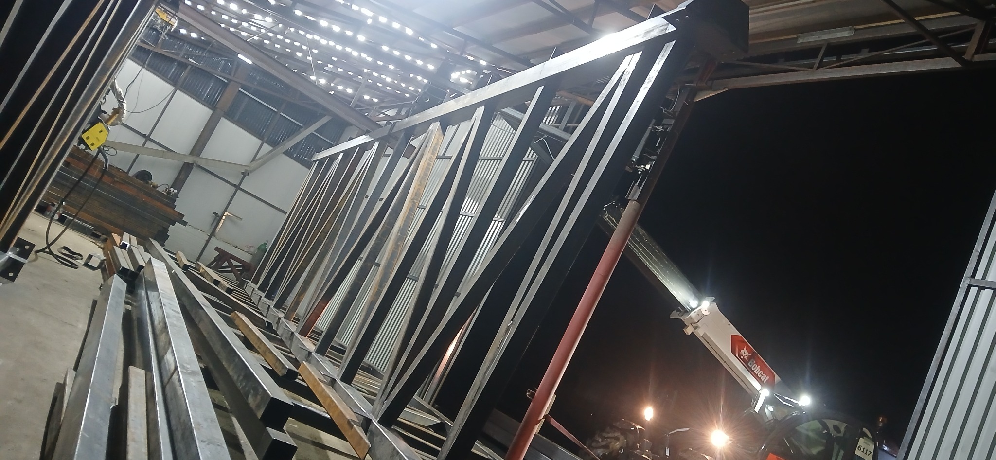 Wnętrze hali produkcyjnej z surową, metalową konstrukcją w trakcie montażu. Widoczny wózek widłowy i elementy spawarki. Nocne oświetlenie.