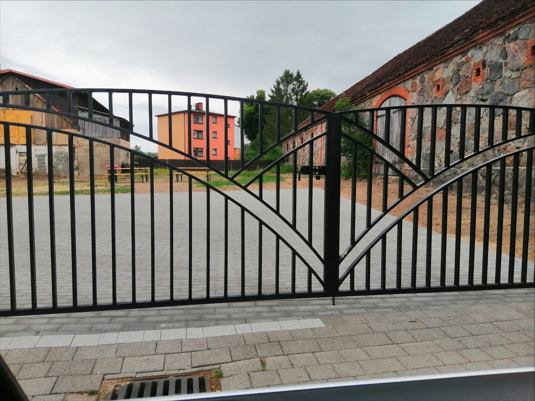 Czarna, metalowa brama wjazdowa z ozdobnymi łukami na tle budynków i szarej kostki brukowej. Widoczny zamek na bramie.